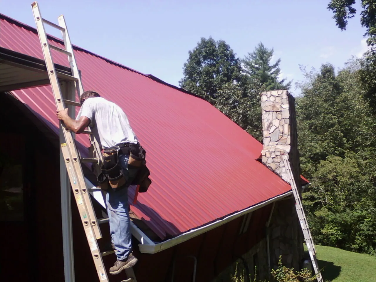 Roofer on a ladder beside a red metal roof and stone chimney — Soffit & Fascia Repair by Roofing Brothers in Fort Hall